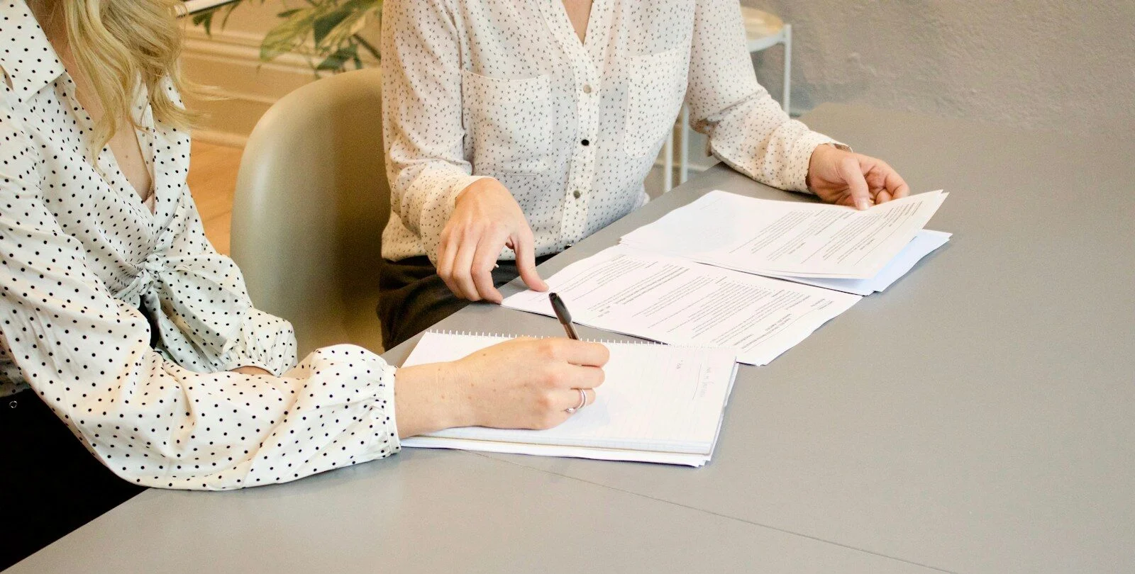 woman signing on white printer paper beside woman about to touch the documents